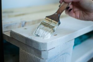 Kobieta skleiła klejem do drewna łóżko z palet i maluje go farbą na biało